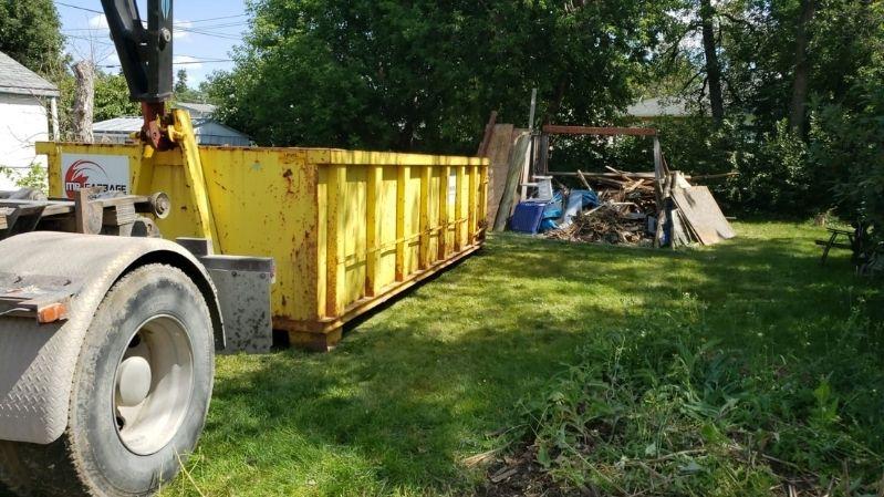 large truck backing up a bin rental into someone's yard. In the distance we see a large pile of garbage